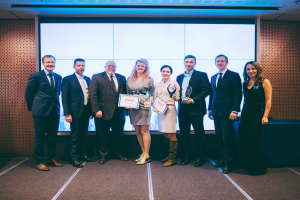 Gruppe von strahlenden Menschen mit Urkunden vor einem Bildschirm, mit einem Tisch und Wandobjekt im Hintergrund, Gewinner der International Business Awards 2018.