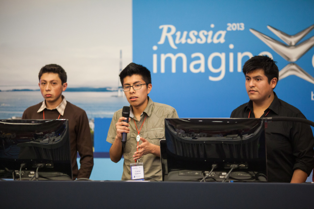 Drei Jungen stehen neben einem Schreibtisch mit zwei Computersystemen, einer spricht in ein Mikrofon und trägt ID-Karten mit roten Markierungen, vor einer Wand mit einer Werbetafel, auf der "Russia Imagine 2013" steht.