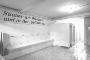 Schwarz-weißes Foto einer öffentlichen Toilette mit Fliesenboden, Wandtext, Wasserhähnen, einer Tür und einer Deckenlampe.