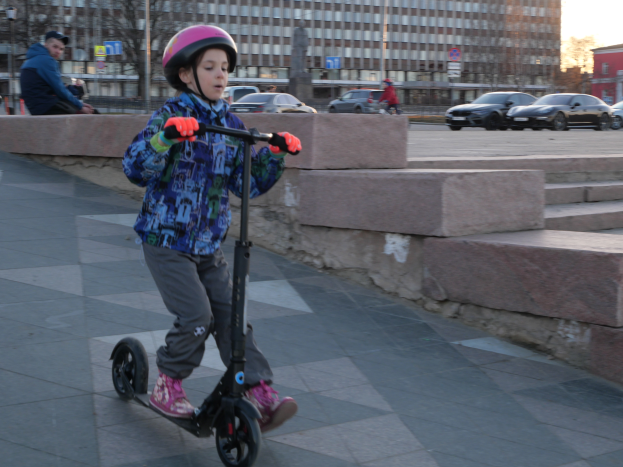 Ein junger Junge fährt mit einem Helm und Handschuhen auf einem Gehweg mit einem Scooter, mit verschiedenen städtischen Elementen und einem klaren blauen Himmel im Hintergrund.