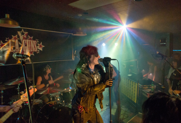 Eine Frau in einer schwarzen Jacke singt in ein Mikrofon auf der B├╝hne, mit einem Mann in Sonnenbrille und einem Headband, der ein Instrument spielt; ein "LA Guns"-Plakat und Diskoleuchten sind im Hintergrund zu sehen.