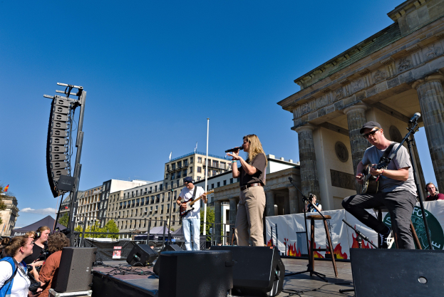 Gruppe von Menschen, die auf einer Bühne mit Instrumenten und Mikrofonen vor dem Brandenburger Tor spielen, mit Lautsprechern und Equipment im klaren blauen Himmel.