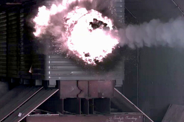 Eine große Explosion in der Mitte eines Gebäudes mit herausquellendem Rauch und einer Wand im Hintergrund sowie metallischen Gegenständen mit einer Tafel unten.