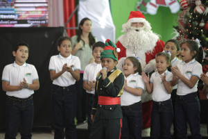 Kinder klatschen neben dem Weihnachtsmann vor einem geschmückten Weihnachtsbaum, wobei ein Kind ein Mikrofon hält und eine Frau im Hintergrund steht.