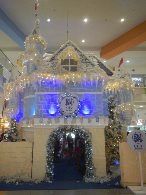 Ein festliches Weihnachtsdorf mit einer burgenähnlichen Struktur, geschmückt mit Lichtern und Schmuck, umgeben von Menschen in einem Einkaufszentrum mit beleuchteter Decke und einem Textschild auf der rechten Seite.
