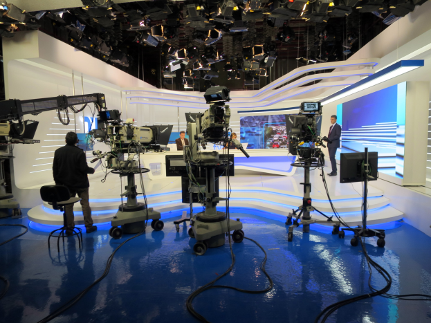 Ein TV-Studio mit mehreren Kameras auf dem Boden, einer sitzenden Person im Vordergrund und einigen stehenden Personen im Hintergrund, ausgestattet mit Deckenleuchten für eine Live-Übertragung.