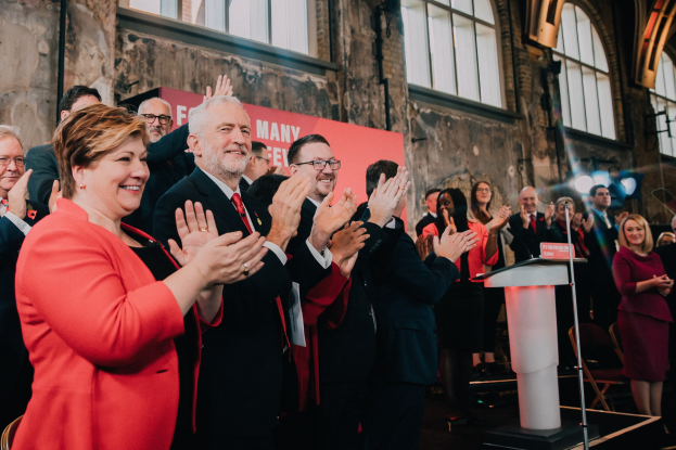 Eine Gruppe von Menschen, die vor einem Publikum jubeln, mit einem Podium, einem Mikrofon und einer Tafel mit Text auf der rechten Seite und Stühlen, einer Fahne, einer Wand, Fenstern und Lichtern im Hintergrund.