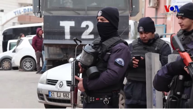Eine Gruppe von Polizisten in Uniformen, die Waffen haltend, steht auf einer Straße in Ankara, Türkei, während einer Anti-Regierungsdemo, mit Fahrzeugen, Menschen, Gebäuden und Texttafeln im Hintergrund.