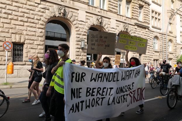 Eine Gruppe von Menschen marschiert auf einer Straße in Berlin, hält Schilder und Banner hoch, während einige Fahrräder fahren, vor einem Gebäude mit Bögen, Säulen, Skulpturen und Bäumen.