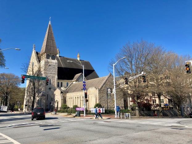 Die Episkopalkirche St. Lukas, eine große Kirche mit einem Turm, steht an einer Straßenecke umgeben von Gebäuden, Verkehrszeichen, Fahrzeugen, Fußgängern und Grünflächen unter einem klaren blauen Himmel.