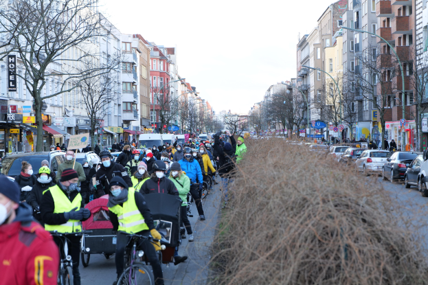 Eine große Gruppe von Menschen in Masken und Sicherheitswesten fährt auf Fahrrädern eine straße mit Bäumen, Gebäuden, Laternen und Texttafeln entlang, während Fahrzeuge die Straße teilen und trockenes Gras die rechte Seite unter einem klaren blauen Himmel einnimmt.
