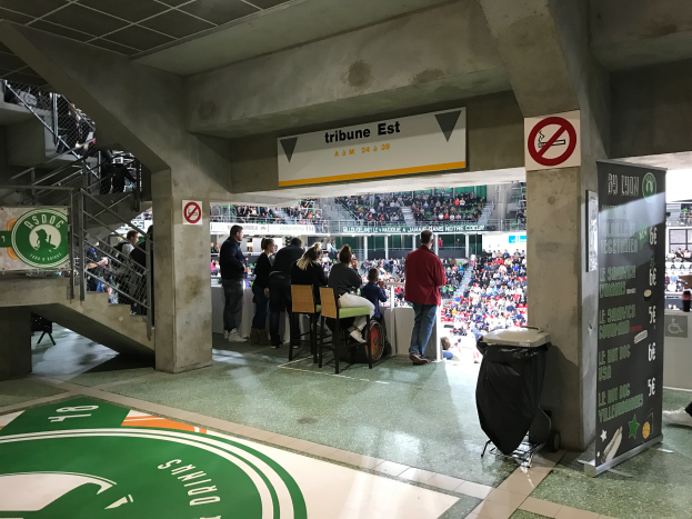 Eine Gruppe von Menschen steht vor einer sitzenden Menge in einem Stadion, mit Schildern, die "Boston Celtics vs New York Jets" lesen, einem Müllcontainer, einer Texttafel, einer Treppe mit Geländern und einer Decke mit Deckenleuchten.