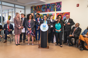 Eine Gruppe von Menschen vor einem Rednerpult mit einem Mikrofon und einer Tafel mit der Aufschrift "Sancho City Councilman Joins City Council", einige sitzen und andere stehen, und ein Hintergrund mit Flaggen, einer Tafel, einem Feuerlöscher und Deckenlampen.