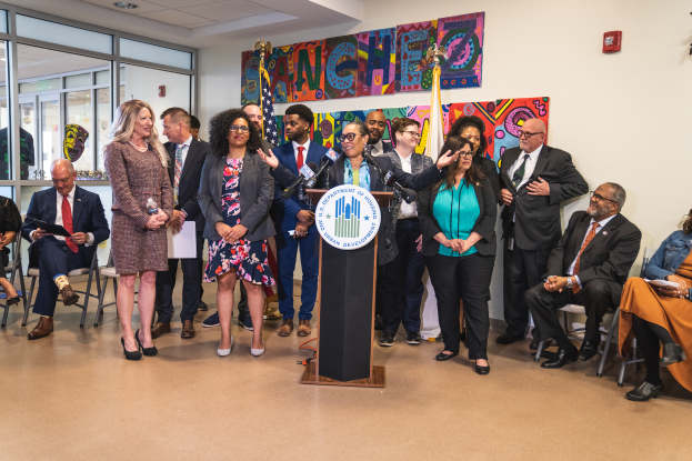 Eine Gruppe von Menschen vor einem Rednerpult mit einem Mikrofon und einer Tafel mit der Aufschrift "Sancho City Councilman Joins City Council", einige sitzen und andere stehen, und ein Hintergrund mit Flaggen, einer Tafel, einem Feuerlöscher und Deckenlampen.