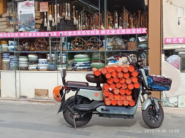 Ein Motorroller mit einem Korb ist auf der Straße vor einem Laden mit einer Glaswand geparkt. Der Laden zeigt verschiedene Gegenstände im Inneren und ein Schild mit Text ist sichtbar.