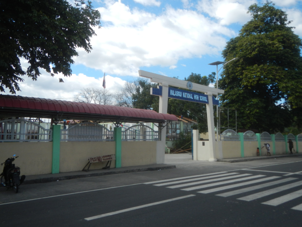 Einfahrt zu einer Schule mit einem vor der Tür geparkten Motorrad, mit einem Tor, einem Schuppen, einem Schild, einer Straße mit weißen Linien und Zebrastreifen, Laternen, Bäumen, einer Fahne und einem bewölkten Himmel.