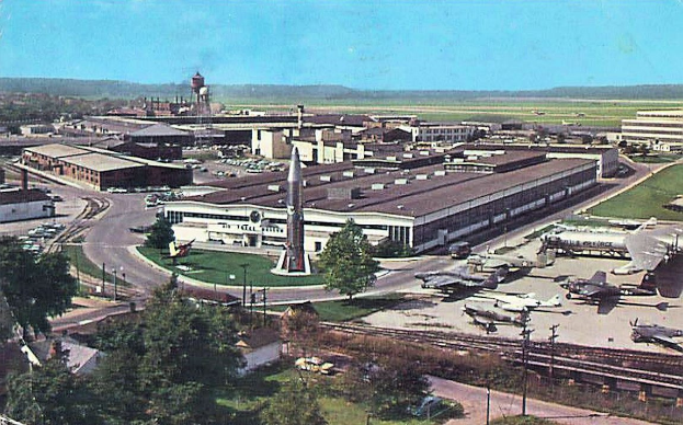 Ein Schwarz-Weiß-Foto eines frühen Flughafens mit geparkten Propellerflugzeugen, Terminalgebäuden, Bäumen, Straßen, Fahrzeugen, Strommasten, Grasflächen, Bahnschienen, fernen Türmen, Hügeln und einem klaren Himmel, mit gedrucktem Text entlang der unteren Kante.