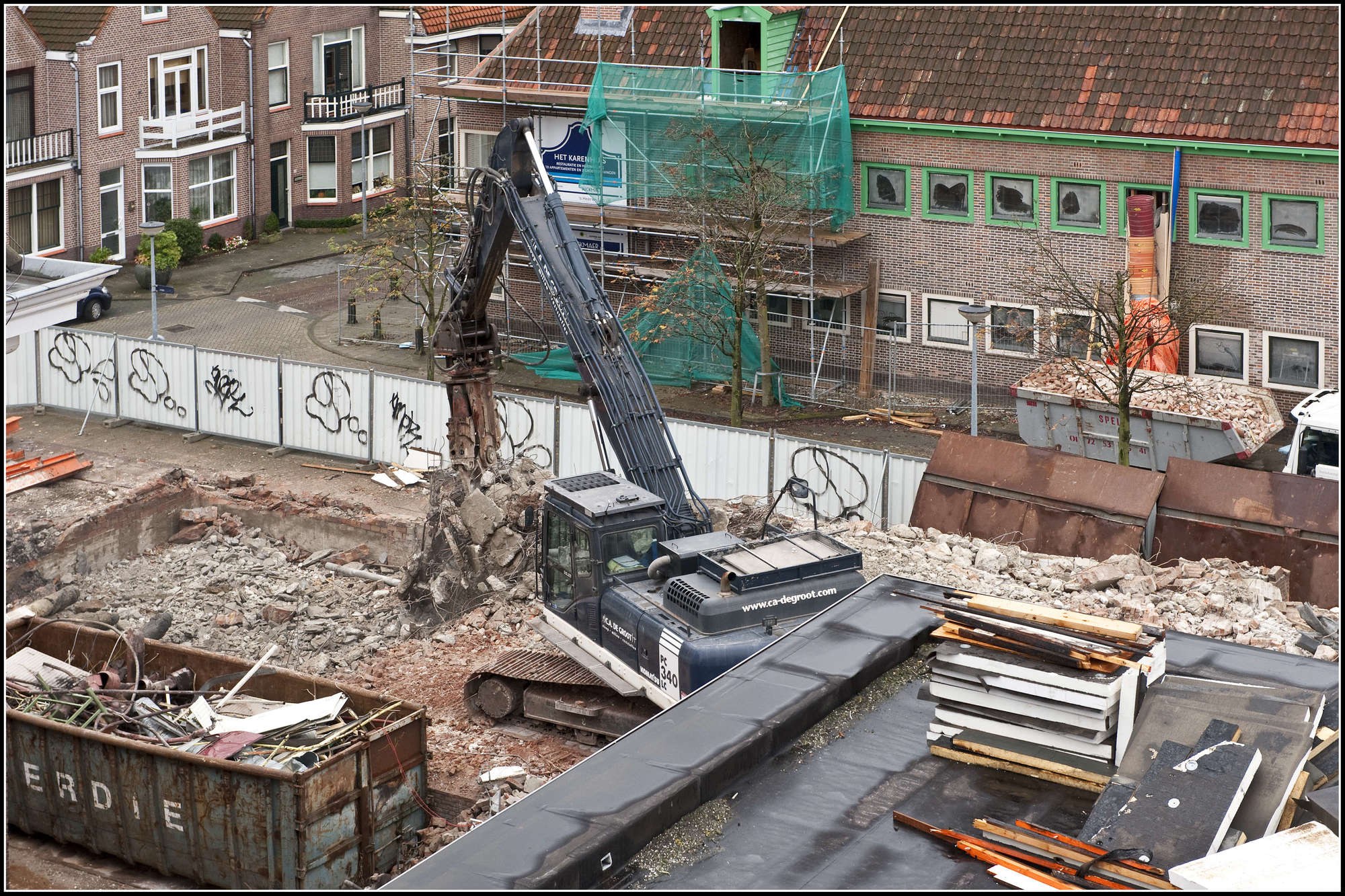 Bagger in Betrieb auf einer Baustelle mit Gebäuden, Bäumen, Fahrzeugen, einem Container und einem Zaun, mit Text am unteren Rand.