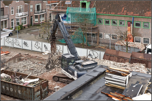 Bagger in Betrieb auf einer Baustelle mit Gebäuden, Bäumen, Fahrzeugen, einem Container und einem Zaun, mit Text am unteren Rand.