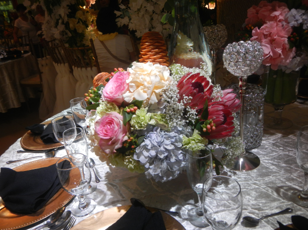 Ein formeller Esstisch mit Tellern, Besteck, Gläsern, Servietten und Blumenvasen, der von einem floralen Hauptgang und Hintergrundstühlen und besetzten Tischen geprägt ist.