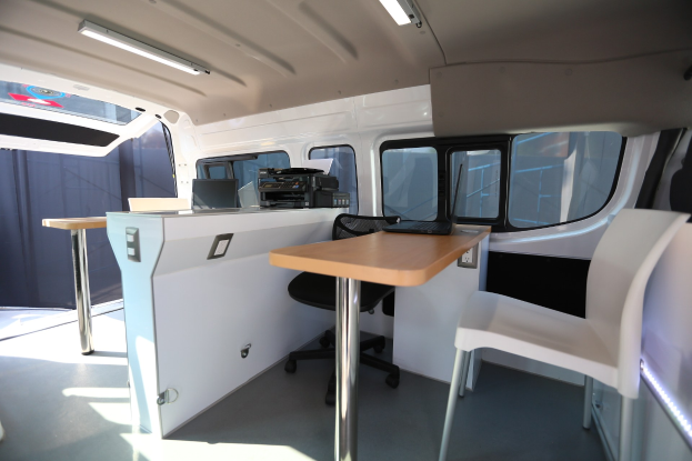Interior of a van with a desk setup, including a laptop, chairs, windows, and overhead lights.