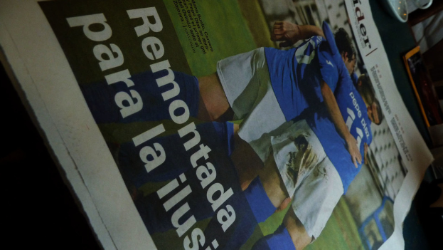 Eine Zeitung auf einem Tisch zeigt drei Männer in blauen Hemden, möglicherweise beim Füßballspielen, mit Text unten auf dem Bild.