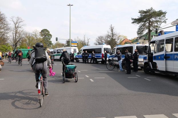 Eine Gruppe von Menschen, die auf Fahrrädern eine Straße entlangfahren, gesäumt von Polizeiwagen und anderen Fahrzeugen, einige tragen Helme und Taschen, andere schieben Kinderwagen, mit Bäumen, Laternenmasten, Verkehrsampeln, Schildern, Gebäuden und einem bewölkten Himmel im Hintergrund.