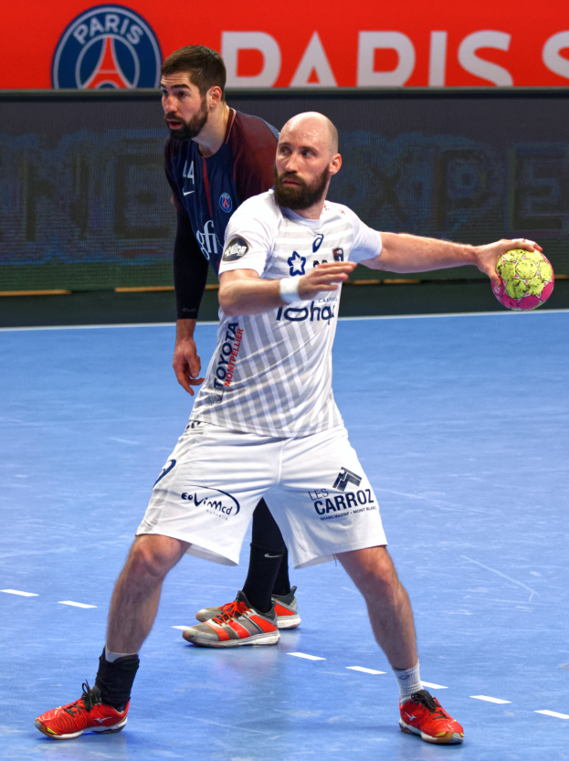 Zwei M├Ąnner beim Handballspielen auf einem Platz, einer h├Ąlt den Ball, mit einer Tafel im Hintergrund, auf der "Paris Saint-Germain vs Paris Saint Germain" steht.