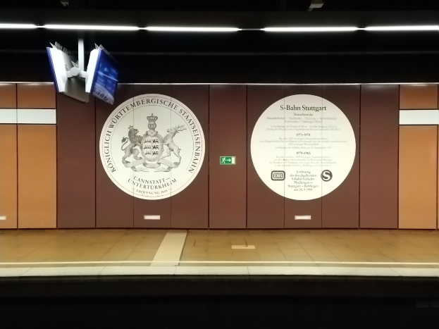 Ein Bahnsteig mit einem Schild "S-Bahn Stuttgart" und einem Logo an der Wand, einige Lampen an der Decke und einige Gegenstände auf der linken Seite.
