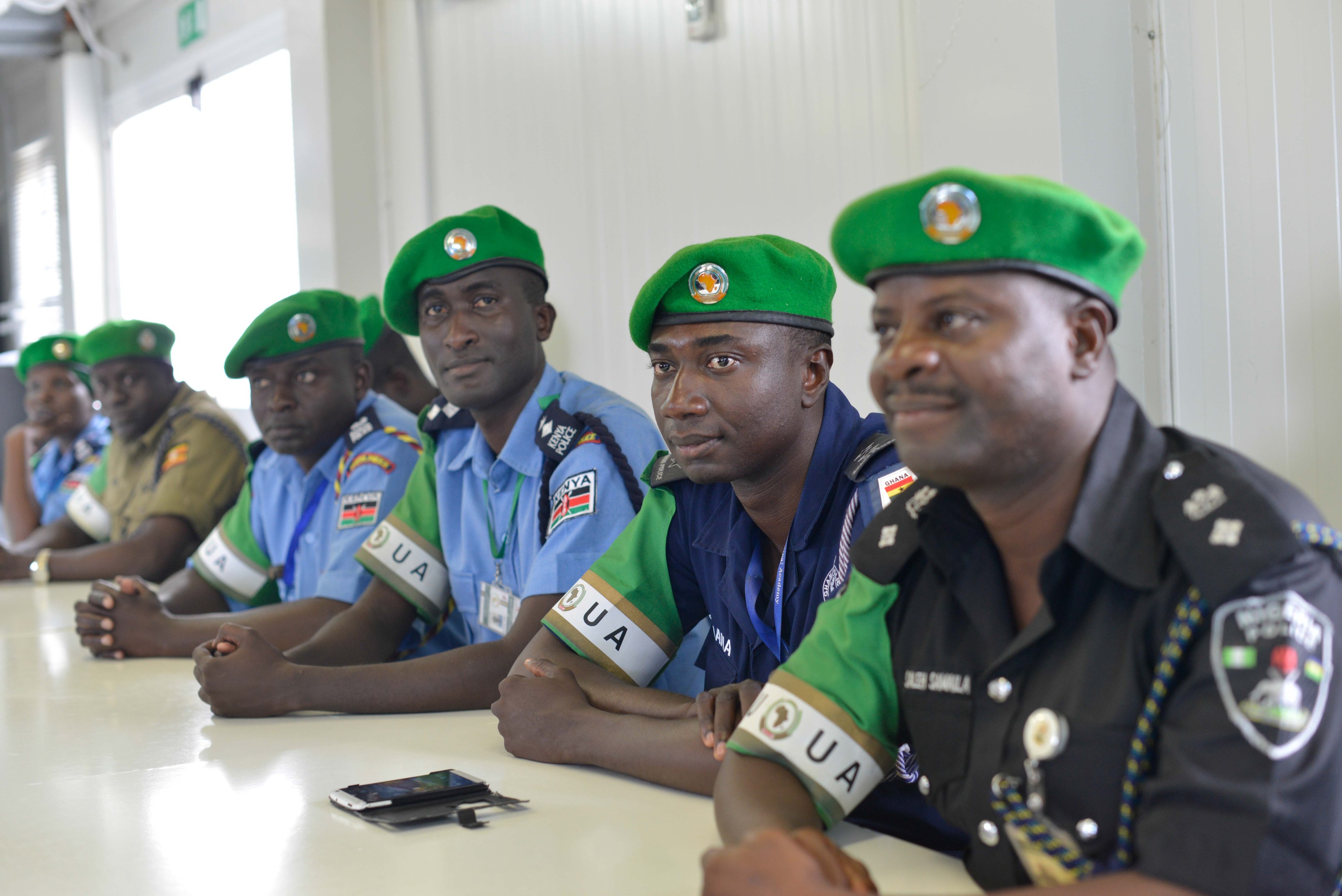 Gruppe von Polizisten, die an einem Tisch während einer Schulung sitzen, mit Mobiltelefonen und anderen Gegenständen auf dem Tisch, in einem Raum mit Fenstern und Deckenbeleuchtung.