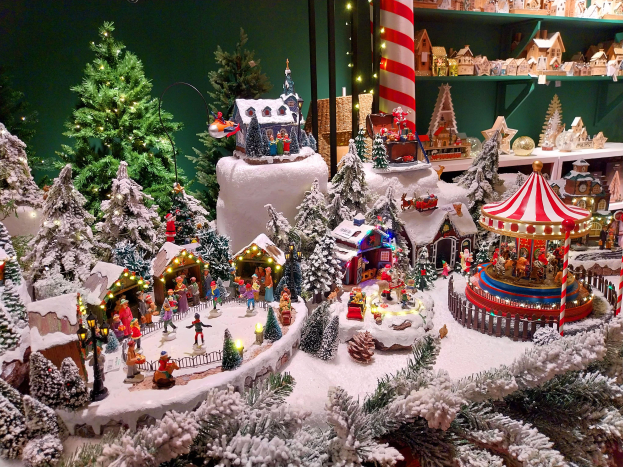 Ein Weihnachtsdorf mit einem Karussell in der Mitte, umgeben von schneebedeckten, beleuchteten Bäumen, Regalen mit Spielzeug auf der rechten Seite und einer Wand im Hintergrund.