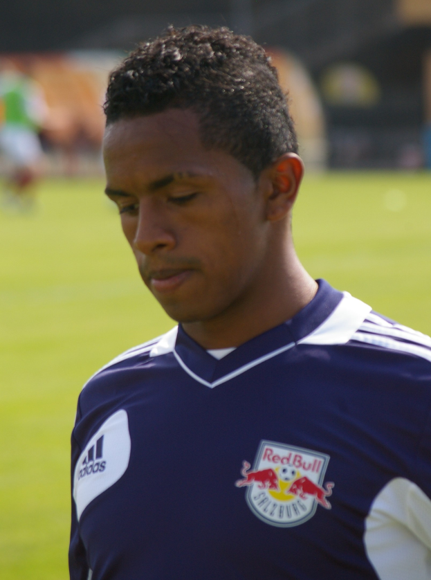 Ein junger Mann in einem blauen T-Shirt, der auf einem Fußballfeld steht, mit einem unscharfen Hintergrund, der wie ein Flügelspieler mit kurzen dunklen Haaren und einem entschlossenen Gesichtsausdruck aussieht.