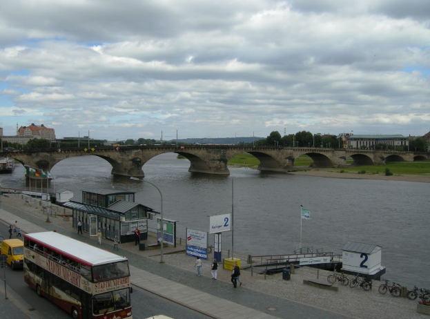 Eine belebte Straßenszene mit Fahrzeugen, Fußgängern, Fahrrädern, Gebäuden, einer Brücke und einem bewölkten Himmel, mit einem Plakat, einer Laterne, Stromleitungen und Gras.