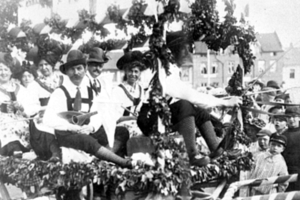 Schwarzes und weißes Foto von Menschen in traditioneller bayrischer Tracht auf einem geschmückten Paradewagen, einige spielen Instrumente, mit Gebäuden und einem klaren Himmel im Hintergrund.