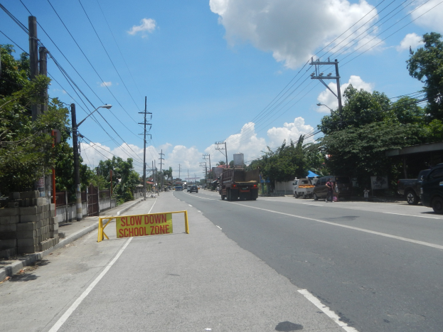 Eine Straße mit Fahrzeugen, ein "Slow Down School Zone"-Schild, Strommäste mit Drähten, Bäume, Laternenpfähle und ein bewölkter Himmel im Hintergrund.