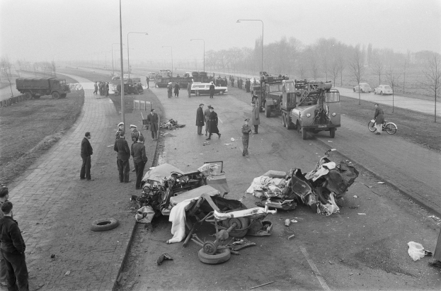 Eine Schwarz-Weiß-Szene eines Autounfalls am Straßenrand mit mehreren Fahrzeugen, darunter ein Landfahrzeug, und einer Gruppe von Menschen drumherum, mit Laternen, Bäumen und Himmel im Hintergrund.