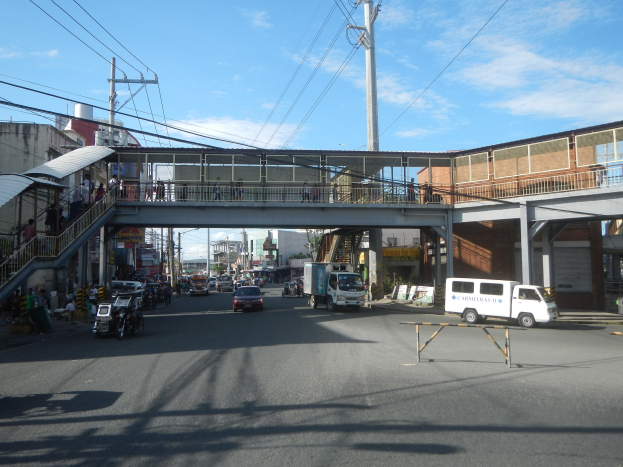 Stadtstraße mit fahrenden Autos, eine Fußgängerbrücke mit Menschen, Strommasten mit Kabeln, Gebäude mit Fenstern und ein bewölkter Himmel.