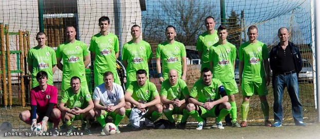 Eine Gruppe von Männern in grünen T-Shirts steht auf einem Fußballfeld, einige hocken, mit einem Ball und einem Torpfosten im Hintergrund; der Hintergrund zeigt Bäume, Gebäude und Fahrzeuge.