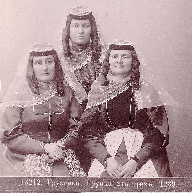 Schwarzes und weißes Foto von drei Frauen in traditionellen russischen Hochzeitskleidern mit farbenfrohen Stickereien und Perlen, die vor einer weißen Wand stehen.