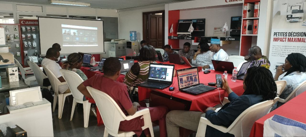 Gruppe von Menschen an einem Tisch mit Laptops und anderen Gegenständen, die an einem digitalen Marketing-Workshop teilnehmen, mit einem Banner, einem Bildschirm und Deckenleuchten im Raum.