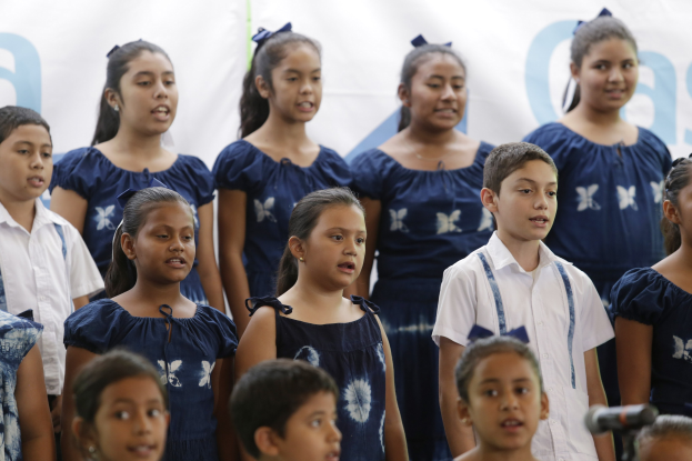Gruppe von Kindern in einem Schulchor, der in ein Mikrofon singt, mit einem Banner im Hintergrund.
