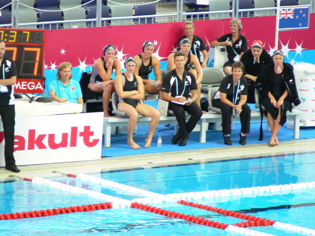 Ein Swimmingpool im Vordergrund mit Personen, die sitzen und stehen, leere Stühle im Hintergrund, eine Flagge, ein Geländer, eine Tafel mit Zahlen und Banner mit Text.