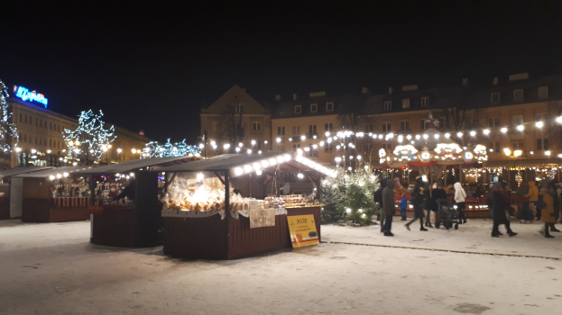 Ein nächtlicher Weihnachtsmarkt in einer verschneiten Straße mit Menschen, Kinderwagen, beleuchteten Buden, Pflanzen, Bäumen, Gebäuden und Schildern unter einem bewölkten Himmel.