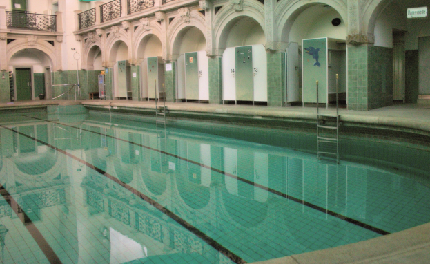 Innenbad mit kristallklarem Wasser, umgeben von gefliestem Boden, in einem großen gewölbten Gebäude mit Säulen, Geländern, Türen und Texttafeln.