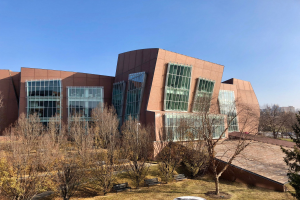 Großes Gebäude mit vielen Fenstern, umgeben von Bäumen, Bänken, Gras und einem Laternenpfahl, mit anderen Gebäuden und einem klaren blauen Himmel im Hintergrund; als der neue Campus der University of Illinois at Chicago identifiziert.