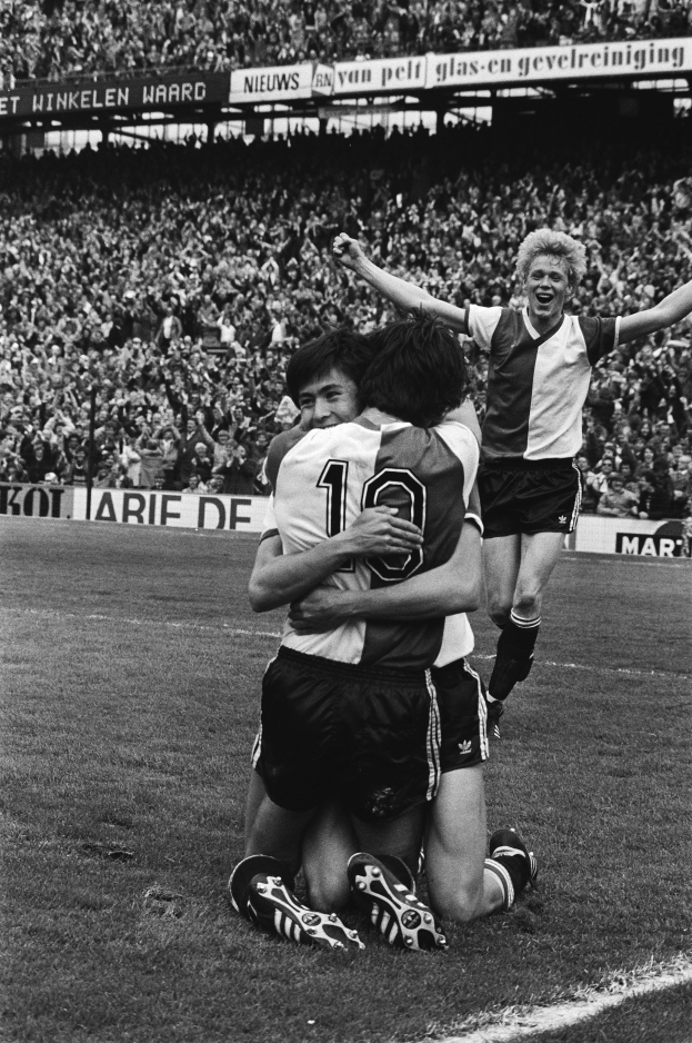 Drei Fussballspieler feiern ein Tor auf dem Boden in einem schwarz-weiss gestalteten Stadion.
