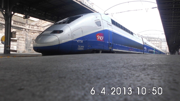 Ein Zug ist in der Mitte des Bildes zu sehen, mit einem Bahnsteig und Text unten, Gebäuden mit Dächern und einer Säule links und Gebäuden mit Dächern, Kabeln und Himmel oben.