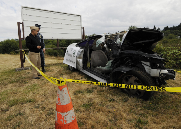 Ein Polizeibeamter in Uniform steht neben einem schwer beschädigten Auto am Straßenrand, mit eingedellter Motorhaube und zersplitterten Fenstern, unter einem bewölkten Himmel mit Bäumen im Hintergrund.