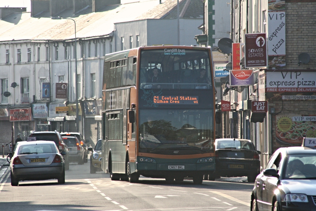 Eine Straße mit Autos und einem Bus im Vordergrund, mit Gebäuden im Hintergrund, die Wände, Fenster, Teller und Dächer aufweisen, sowie mit Plakaten und Bannern geschmückt sind, und ein Laternenmast.