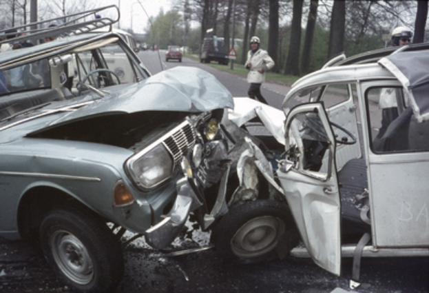 Ein verunfalltes Auto am Straßenrand, umgeben von anderen Fahrzeugen und Menschen mit Helmen, mit Bäumen, Pfählen und einem klaren blauen Himmel im Hintergrund.